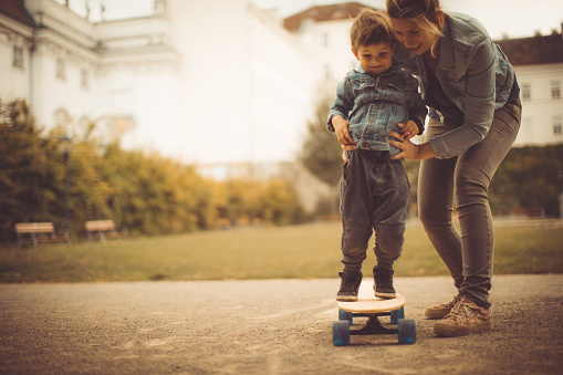 Little smiling boy and his mother are learning how to skate in the park on skateboard 