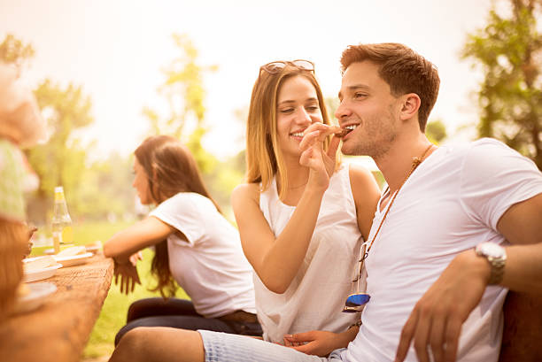 Young people having fun during barbecue picnic in the park or back yard. Woman in front view is feeding her boyfriend with roasted meat. Food and drink are on the wooden table.