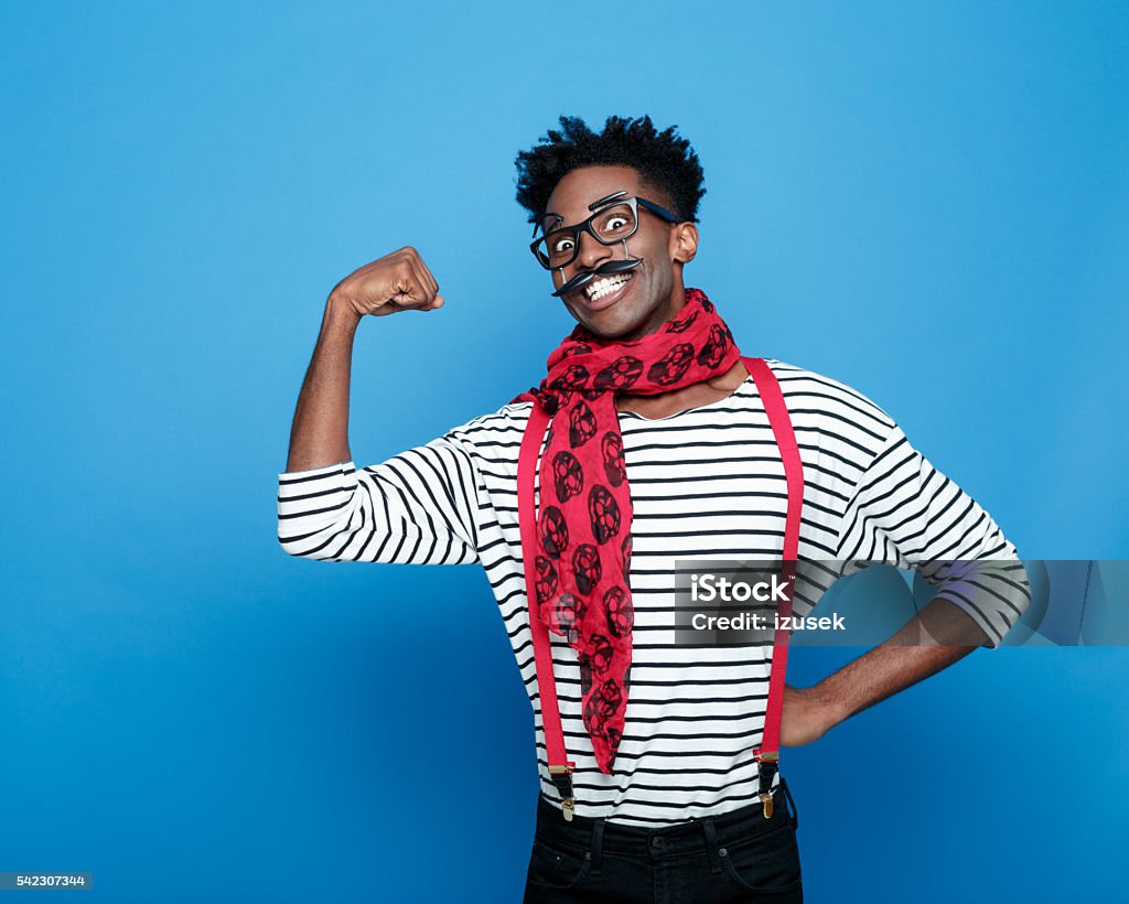 Excited afro american guy in a french outfit flexing bicep Portrait of excited afro american guy wearing striped long sleeved t-shirt, red suspenders and neckscarf, flexing his bicep and grining at the camera. Studio shot, blue background.  Bizarre Stock Photo
