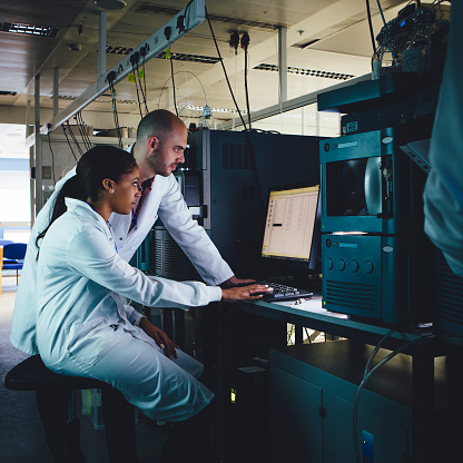 Male and female scientists discussing data on a computer screen amongst large medical machinery.