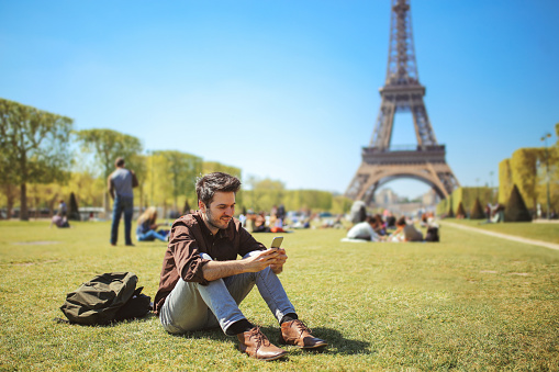 Vintage toned image of a young man relaxing, using his cellphone, having a break near the Eiffel Tower park on a bright and sunny day in Paris, France. He is wearing a smart casual outfit, brow leather shoes and a shirt, having his backpack by. Modern lifestyle, casual fashion concepts.