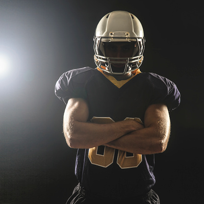 Back lit American football player with crossed arms. Unrecognizable male on black background.