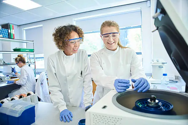 a female researcher is standing in the foreground at her lab bench  transferring genetic samples to her centrifuge being supervised by a colleague or a teacher who is explaining the process. In the background a female colleague is sitting at her bench pipetting samples to a microplate