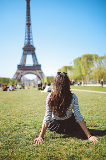 Vintage toned image of a young woman relaxing in the park near the Eiffel Tower on a bright and sunny day in Paris, France. She is wearing a casual, street style outfit, a striped shirt and a skirt. Modern lifestyle, casual fashion, relaxation in the summertime concepts.