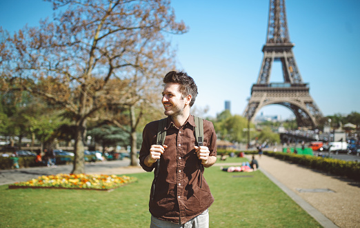 Vintage toned image of a young tourist, walking and relaxing relaxing in the park near the Eiffel Tower on a bright and sunny day in Paris, France. He is wearing a smart casual outfit, brow leather shoes and a shirt, having his backpack by. Modern lifestyle, casual fashion concepts.