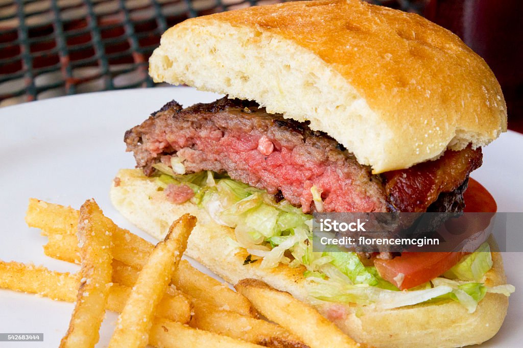 Delicious hamburger with fried potatoes Grilled hamburger with bacon, shredded lettuce, tomatoe slice.  Meat on bread roll with fried potatoes on outdoor, wrought iron patio table. Rare - Cooking Technique Stock Photo