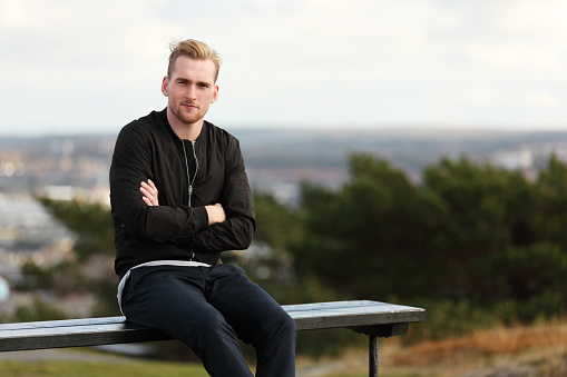 An attractive man in his 20s wearing a black jacket sitting down outside on a windy autumn day looking away from camera. 