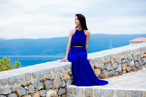 photo of the beautiful young woman sitting on the stone railing and looking at the splendid view in Greece