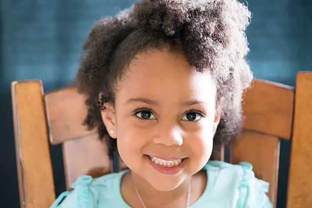 African American young girl/child with natural hair pose sitting with eyes looking upwards to the camera with contrast lighting where her face is in shadow and side hair in light with a smile.