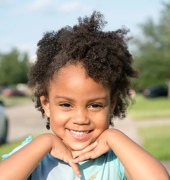 African American young girl/child with natural hair standing looking forward with fingers crossed interlocked under chin and smiling.