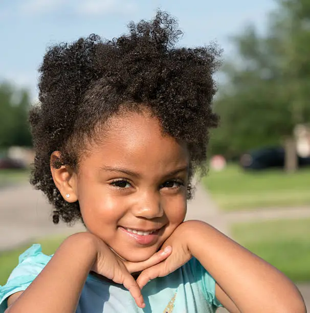 African American young girl/child with natural hair standing looking forward with fingers crossed interlocked under chin with head slightly turned towards the camera.
