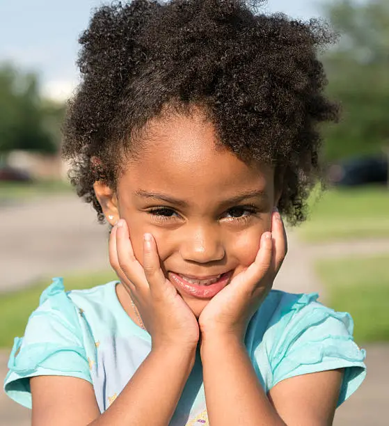 African American young girl/child with natural hair standing looking forward with hands under chin with a slight smile.