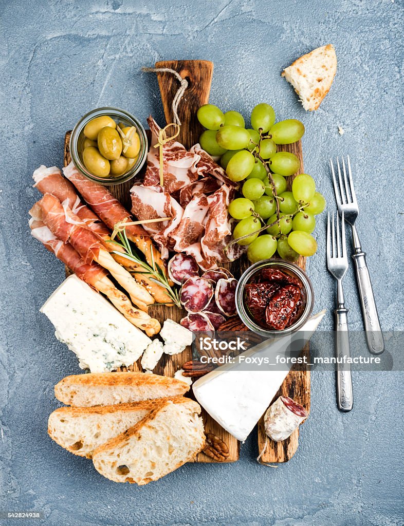 Cheese and meat appetizer selection. Prosciutto di Parma, salami, bread Cheese and meat appetizer selection. Prosciutto di Parma, salami, bread sticks, baguette slices, olives, sun-dried tomatoes, grapes and nutson rustic wooden board over grey concrete textured backdrop, top view Plank - Timber Stock Photo