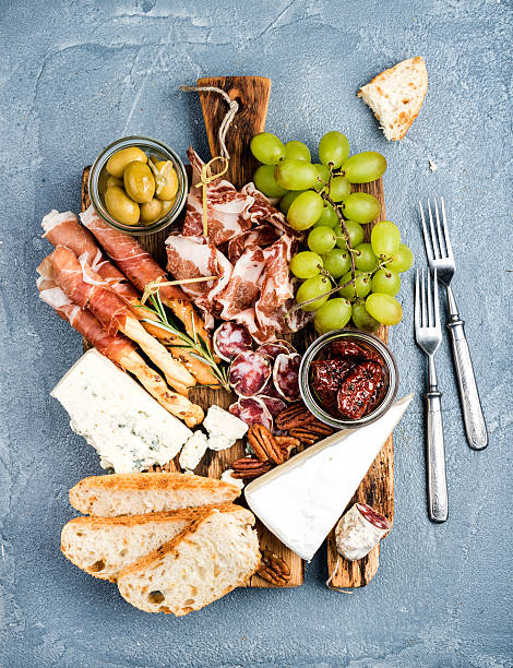 Cheese and meat appetizer selection. Prosciutto di Parma, salami, bread Cheese and meat appetizer selection. Prosciutto di Parma, salami, bread sticks, baguette slices, olives, sun-dried tomatoes, grapes and nutson rustic wooden board over grey concrete textured backdrop, top view salumeria stock pictures, royalty-free photos & images