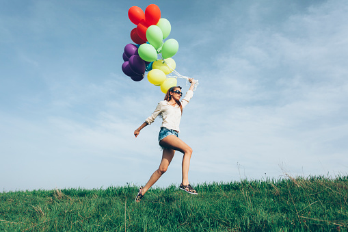 Side view of cheerful young woman holding a bunch of colorful balloons and running, hand raised. Wears casual clothes and eyeglasses.