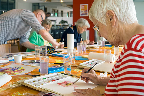 Woman working on watercolor painting. Woman in striped red and white shirt working on watercolor painting at table with other students in spacious studio. activity stock pictures, royalty-free photos & images