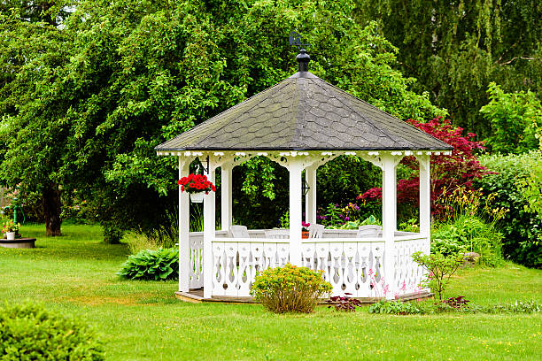 White gazebo Lovely white gazebo with red flowers hanging from a basket. Trees and shrubs in the background. Fine garden with green grass. pavilion photos stock pictures, royalty-free photos & images