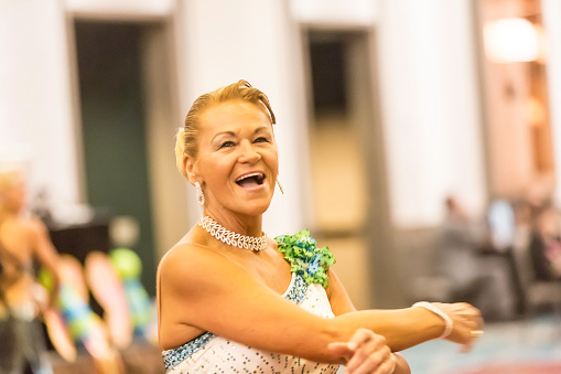 In this real situation a caucasian senior ballroom dancer is taking part in a ballroom dance competition. She dressed in ballroom dancing clothes. she is wearing a purple white dress with sequins. Her partner is not in the picture. She is smiling at him. Taken with Canon 5D Mark 3.