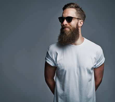 Portrait of handsome single bearded young man with serious expression wearing sunglasses and white short sleeve shirt looking over gray background with copy space