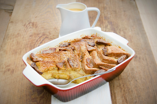 Traditional British Dessert, Bread and Butter Pudding with Custard. Served in a red oven dish on wood. 