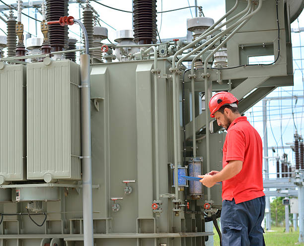 Electrician and High Voltage Transformer Engineer with red hardhat and protective workwear at work in power plant, near transformer. electricity transformer photos stock pictures, royalty-free photos & images