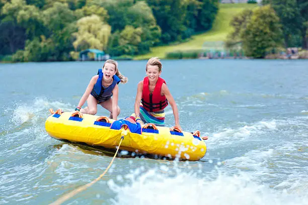 Boy and girl, brother and sister, two active siblings tubing together across a lake in Minnesota, USA. The high speed water sport on an inflatable raft is a family summer vacation activity at Midwest tourist destinations. The children smile playfully as they float across the water surface, flying over waves. 