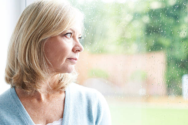 triste mujer madura que sufre de agorafobia mirando fuera de windo - women depression window sadness fotografías e imágenes de stock