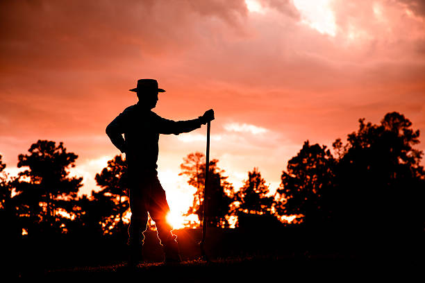 Cowboy, rancher, or farmer overlooking his crops, field after a rain storm at sunset. He is using a shovel as he stands silently on a hill.  Dramatic sky.  Pine tree.   Silhouette.  Lens flare.