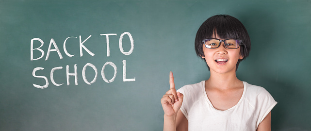 asian girl standing in front of chalk board front view with one finger up laughing with back to school in the blackboard