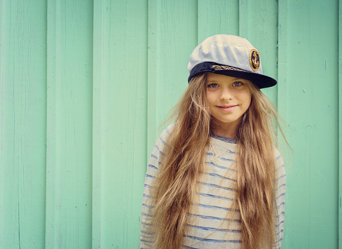 Cute little girl stands near a turquoise wall in sailor hat and smiling Space for text. Negative space.