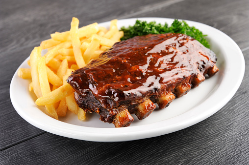 Grilled juicy barbecue pork ribs in a white plate with fries and parsley.
