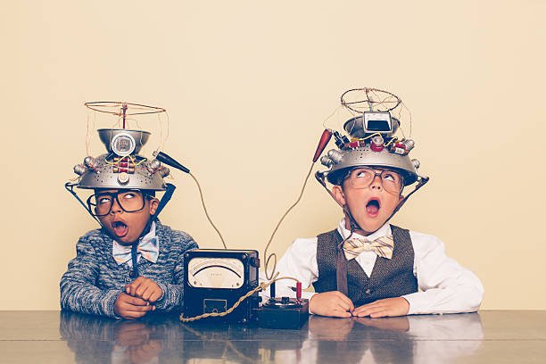 Two Boys Dressed as Nerds Experimenting with Mind Reading Helmets Two nerd boys dressed in casual clothing, glasses and bow ties experiment with a homemade science project. They are sitting at a table with stressed expressions with helmets on their heads in front of a beige background. Retro styling. stem topic stock pictures, royalty-free photos & images