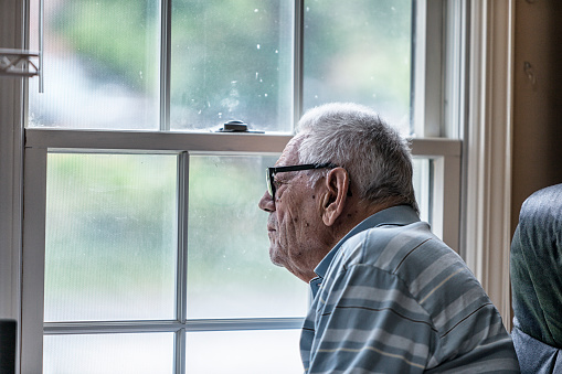 A senior man with early dementia is wearing a hearing aid. He is sitting and staring through the mesh screen and spotted, speckled, unwashed windows at his home.