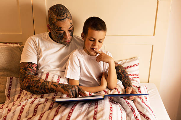 giovane padre leggendo un libro un suo figlio in camera da letto. - tattoo father family son foto e immagini stock