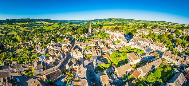 Aerial view over the iconic Cotswold village of Painswick, with its honey coloured limestone cottages and historic church spire, Gloucestershire, UK, framed by vibrant green patchwork fields and clear blue summer skies. ProPhoto RGB profile for maximum color fidelity and gamut.
