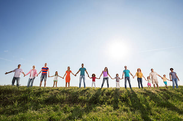 Large group of happy people holding hands against the sky. Low angle view of group of smiling people standing in a meadow against the sky and holding hands while looking at camera. Copy space. line of people holding hands stock pictures, royalty-free photos & images