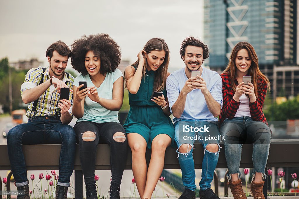 Connecting and sharing Multi-ethnic group of people sitting on an urban bridge text messaging and sharing smart phone content. Friendship Stock Photo