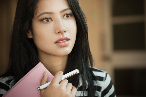 Indoor, day time, close-up image of a serene Asian teenager girl student with long and black hair. She is thinking and looking away at something with blank expression on her face holding a book and pen. One person, head and shoulders, horizontal composition with copy space and selective focus.