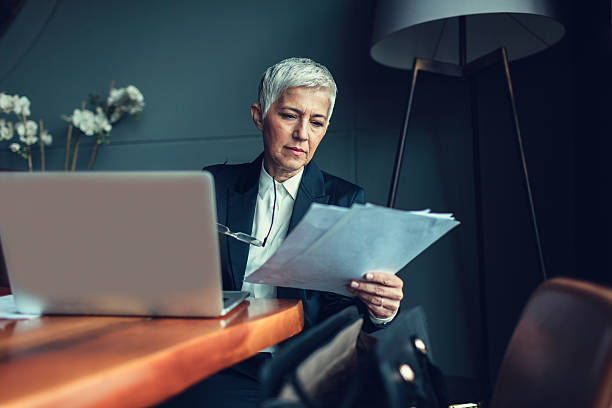 Mature Businesswoman In Her Office. Portrait of mature businesswoman, sitting in her office and finishing paperwork. Holding eyeglasses. In front of her on the desk is laptop. leanincollection stock pictures, royalty-free photos & images
