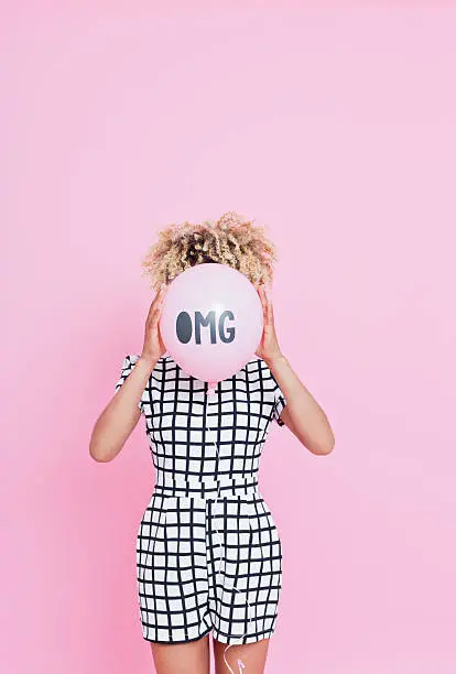 Portrait of unrecognisable young woman, wearing grid check playsuit, standing against pink background and hide her face behind OMG Balloon.