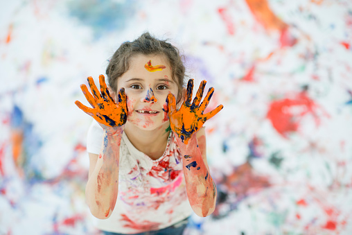 A little girl is creatively playing with finger paints for art therapy. The girl and the white background is covered with hand prints, paint splatters, and colorful paint.