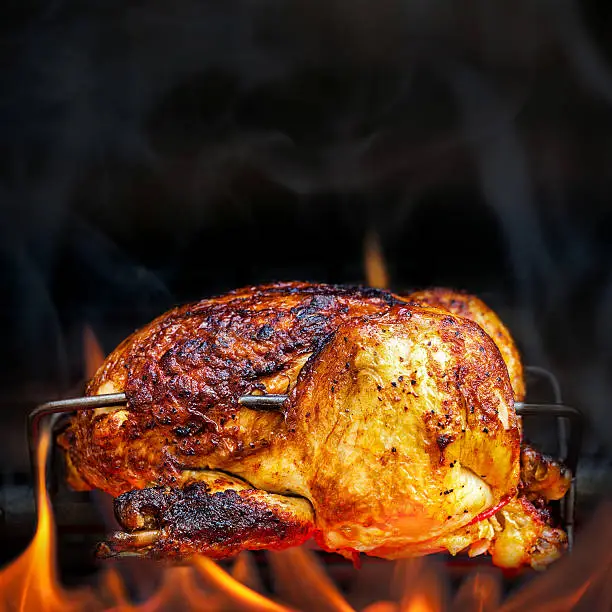 Charred rotisserie chicken over open flames in a barbecue. Square format with room for text