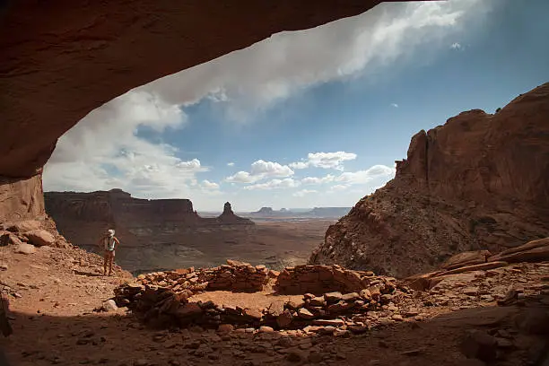 Under a protective alcove, a woman hiker looks out over the desert panorama near an ancient puebloan stone shelter as stormy rain clouds descend over the mesas and spires of the "Islands of the Sky" portion of Canyonlands National Park in Utah.