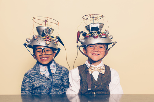 Two nerd boys dressed in casual clothing, glasses and bow ties experiment with a homemade science project. They are smiling and sitting at a table with helmets on their heads in front of a beige background. Retro styling.