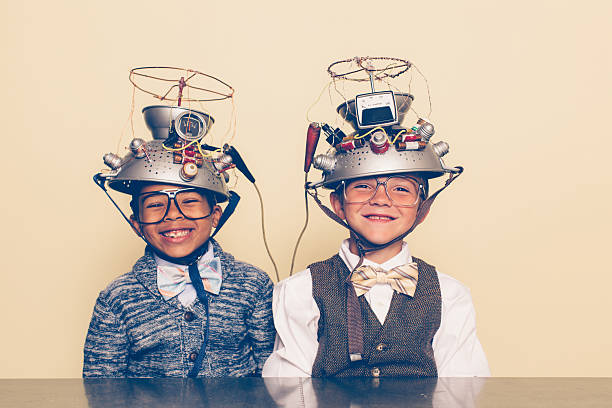 Two Boys Dressed as Nerds Smiling with Mind Reading Helmets Two nerd boys dressed in casual clothing, glasses and bow ties experiment with a homemade science project. They are smiling and sitting at a table with helmets on their heads in front of a beige background. Retro styling. male friendship stock pictures, royalty-free photos & images