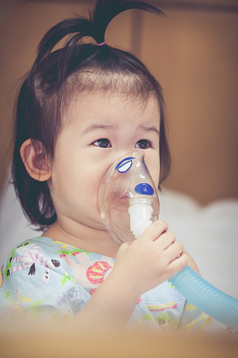 Cute asian child holds a mask vapor inhaler for treatment of asthma on sickbed in hospital. Breathing through a steam nebulizer. Concept of inhalation therapy apparatus. Vintage tone.