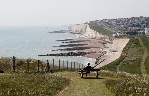 These are the white chalk cliffs of southern England photographed from Rottingdean and looking towards Brighton. In the other direction the cliffs run down to Dover in Kent. The concrete extensions built out from the beach are there to control coastal erosion. These battlements are called groynes. Seated on a bench, looking out to sea, is a Russian outdoor girl model.