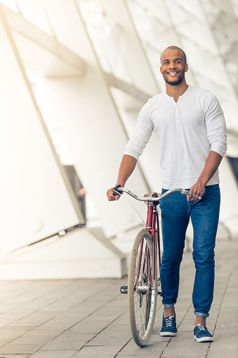 Handsome young Afro American man in casual clothes is smiling while walking with bike outdoors
