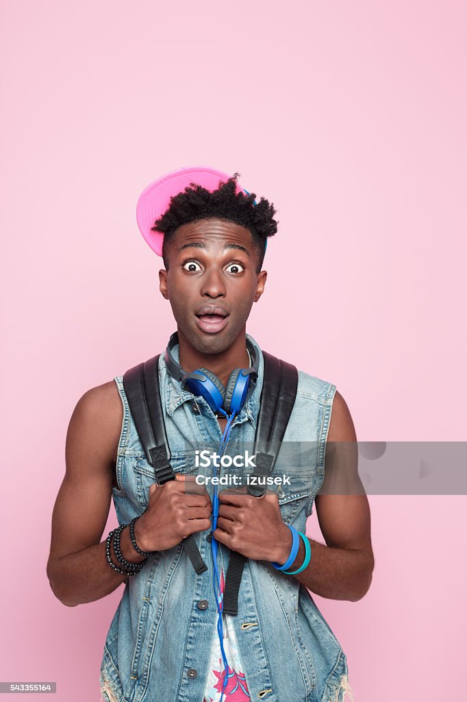 Summer portrait of surprised afro american guy Summer portrait of surprised afro american young man wearing headphone, cap and jeans sleeveless jacket, standing against pink background, staring at camera with mouth open. African Ethnicity Stock Photo