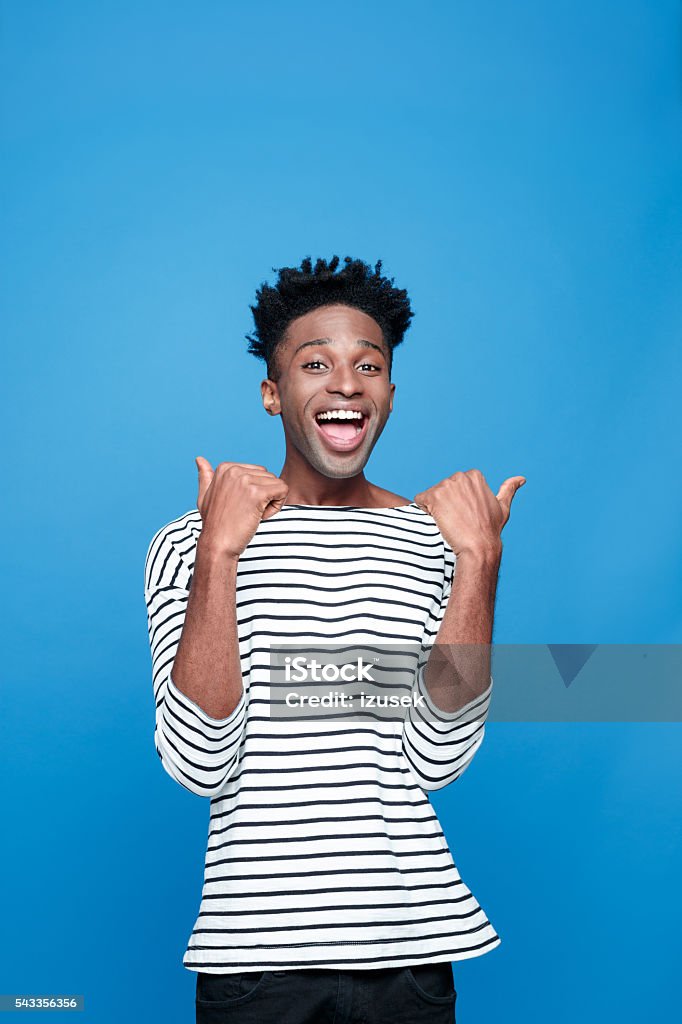 Excited afro american guy Portrait of happy afro american young man wearing striped top, laughing at camera with thumbs up. Studio portrait, blue background. Happiness Stock Photo